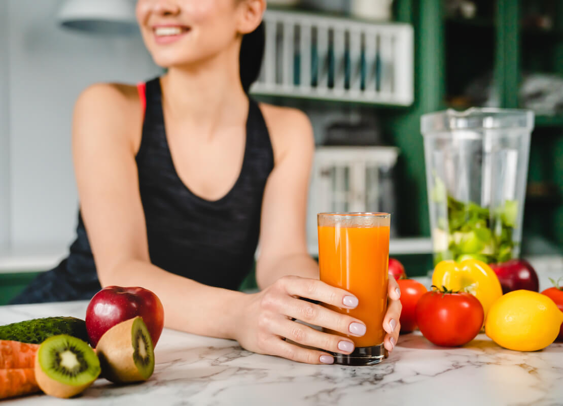Close up of womans hands around a smoothie in a clear glass resting on kitchen counter with colorful fruits and vegetables sitting nearby