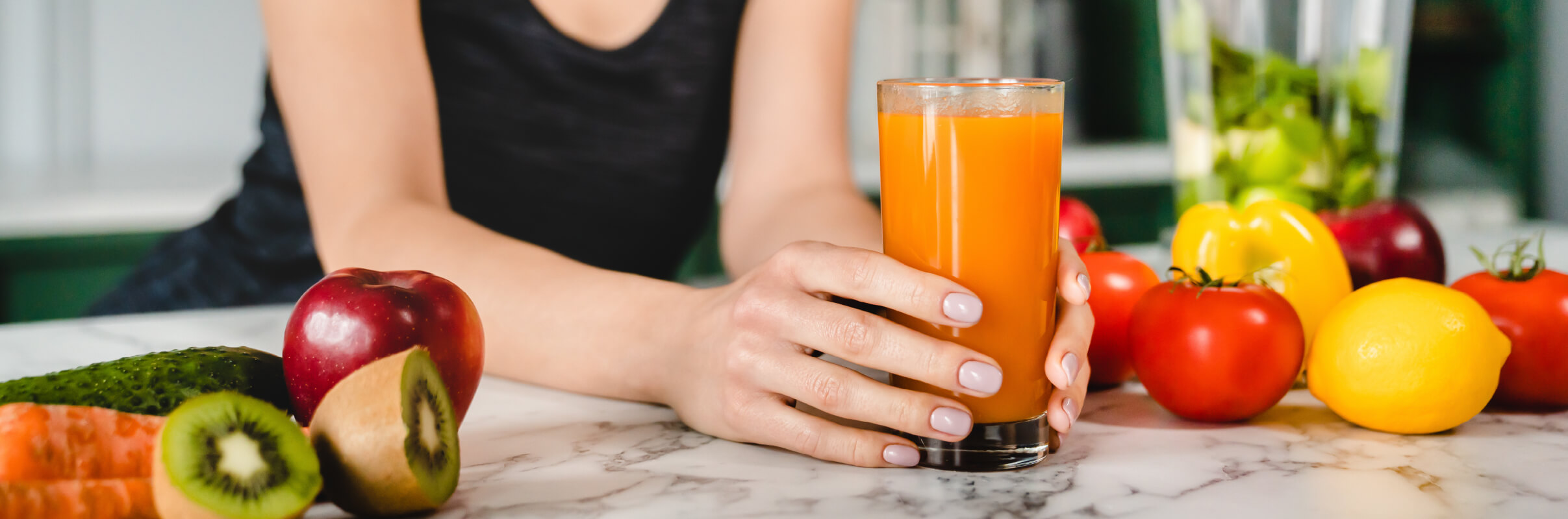 Close up of womans hands around a smoothie in a clear glass resting on kitchen counter with colorful fruits and vegetables sitting nearby